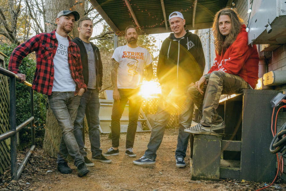 Bandfoto: Die fünf Bandmitglieder stehen nebeneinander und nett lächelnd da. Sie befinden sich draußen, aber unter einem Dach. Hinter ihnen blitzt die Sonne durch