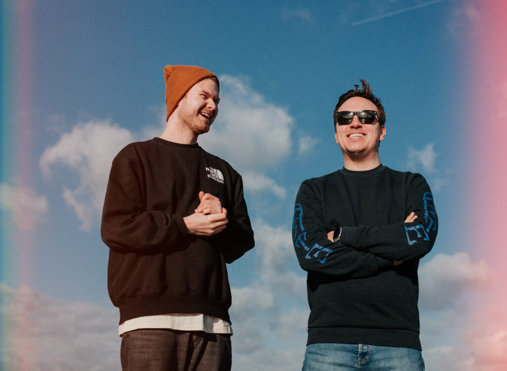 Die beiden Bandmitglieder stehen lachend im Zentrum des Bildes. Hinter ihnen sehen wir einen blauen Himmel mit ein paar weißen Wolken.