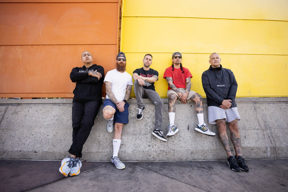 Band Foto: Die fünf Mitglieder sitzen auf bzw. lehnen an einer Mauer. Die Wand im Hintergrund ist auf der linken Hälfte orange und auf der rechten gelb. Die Band wurde von schräg von unten fotografiert, sodass man sie komplett sehen kann.